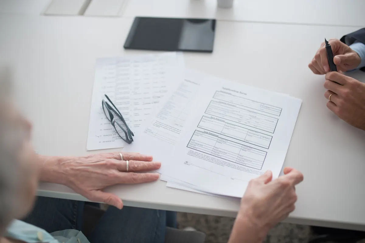 An older person's hands resting on a printed form and papers on a table, with glasses and a tablet nearby