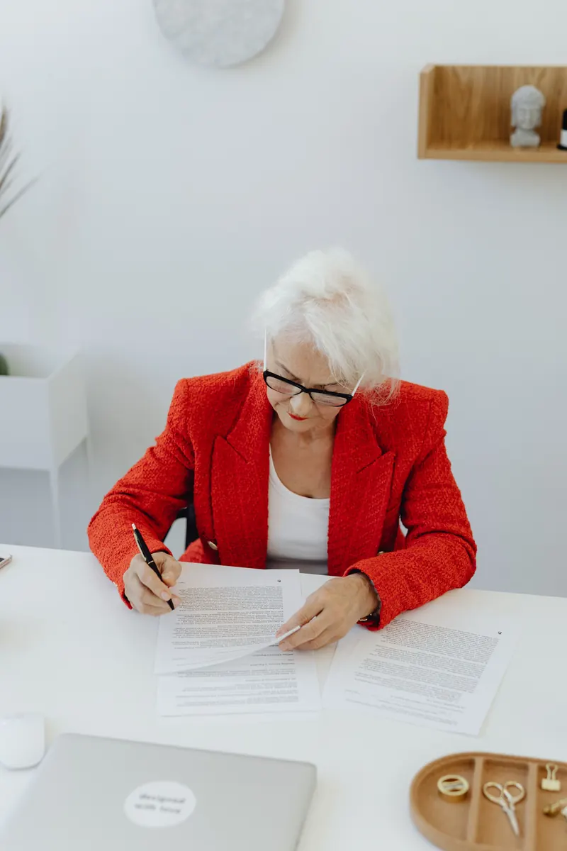 A woman with white hair and glasses in a red blazer reading and marking printed papers at a white desk