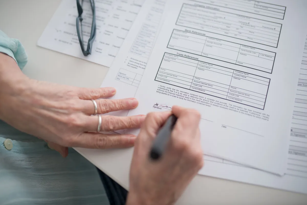 An older person's hands signing a printed form on a table, with reading glasses on papers nearby