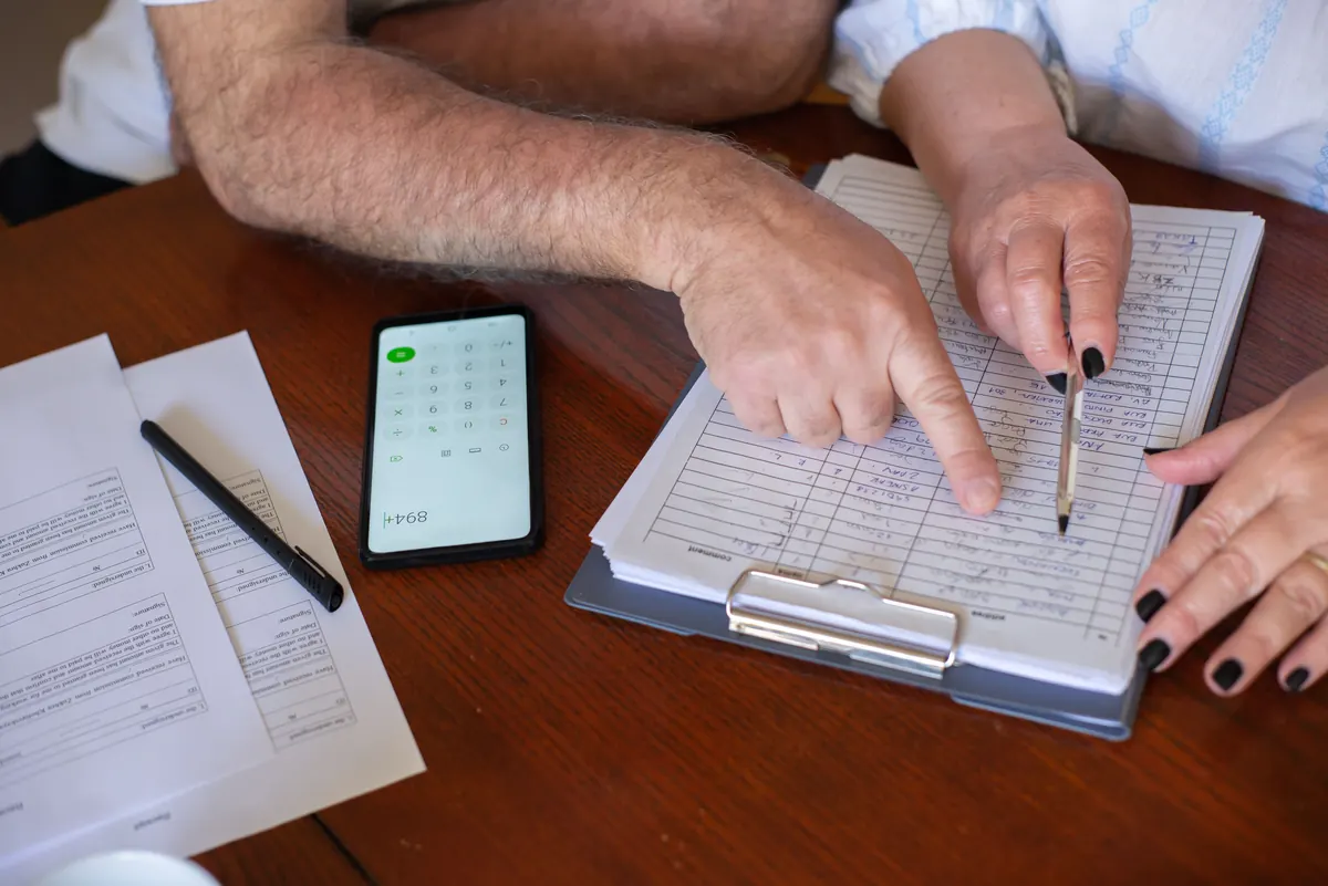 Two people's hands going over a handwritten list on a clipboard, with a phone calculator and forms nearby