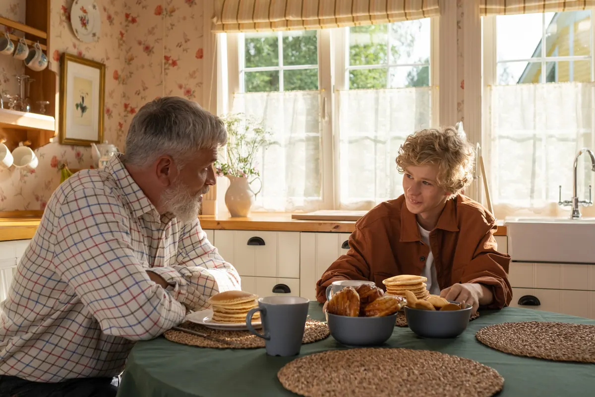 An older man with a gray beard and a teenage boy talking over pancakes at a sunny kitchen table