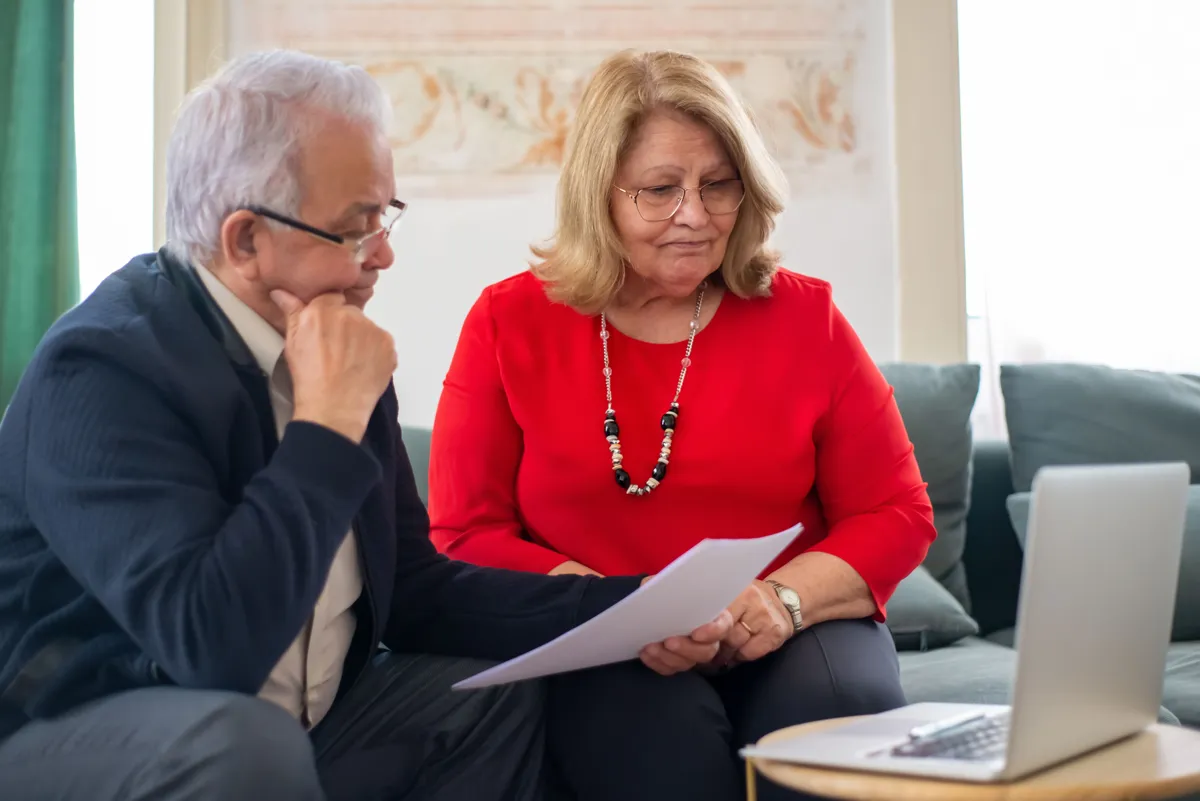 An older couple reading a paper together on their sofa, the woman in a red top