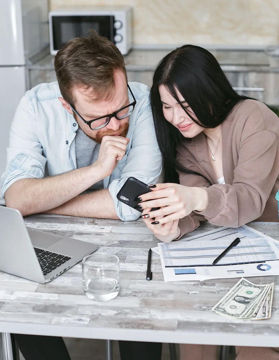 A man and a woman at a kitchen table looking at a phone, with a laptop, papers and cash in front of them