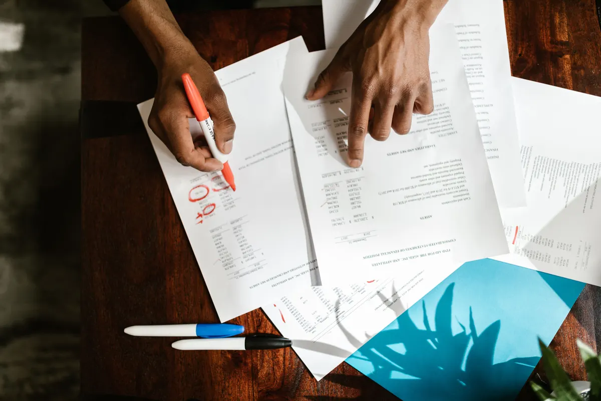 Two hands marking printed statements with an orange marker on a wooden table, seen from above