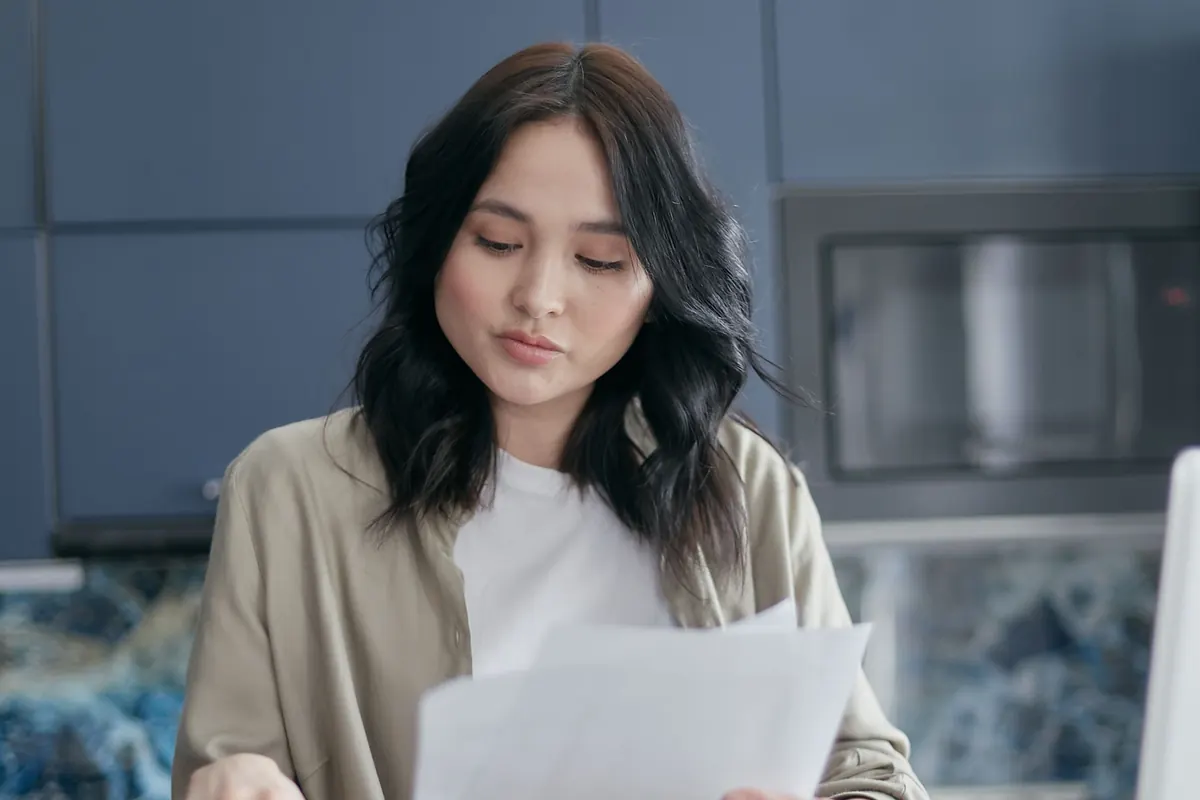 A young woman with long dark hair reading printed papers in a kitchen