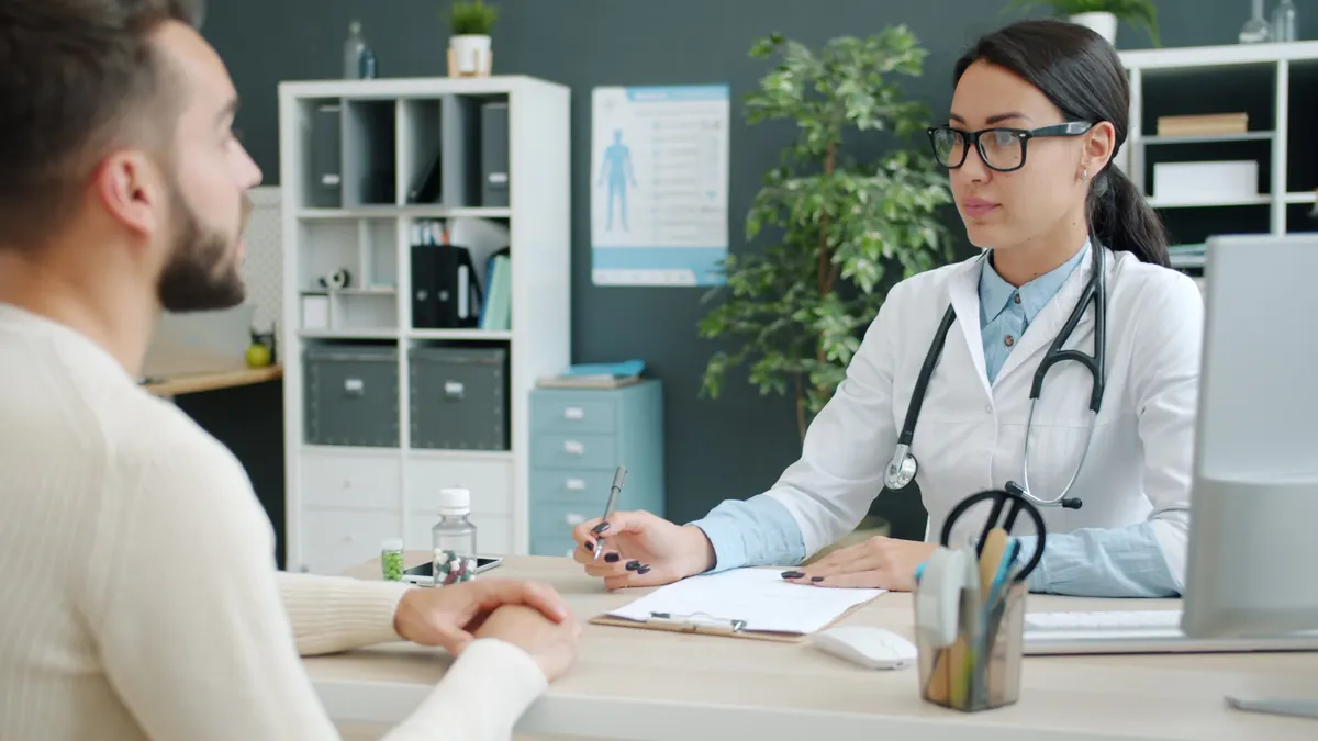 A doctor in a white coat with a stethoscope talking with a bearded man across a desk in her office