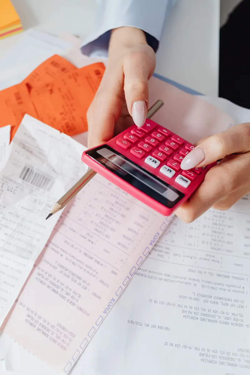 Hands holding a pink calculator and a pencil over a pile of printed receipts and invoices