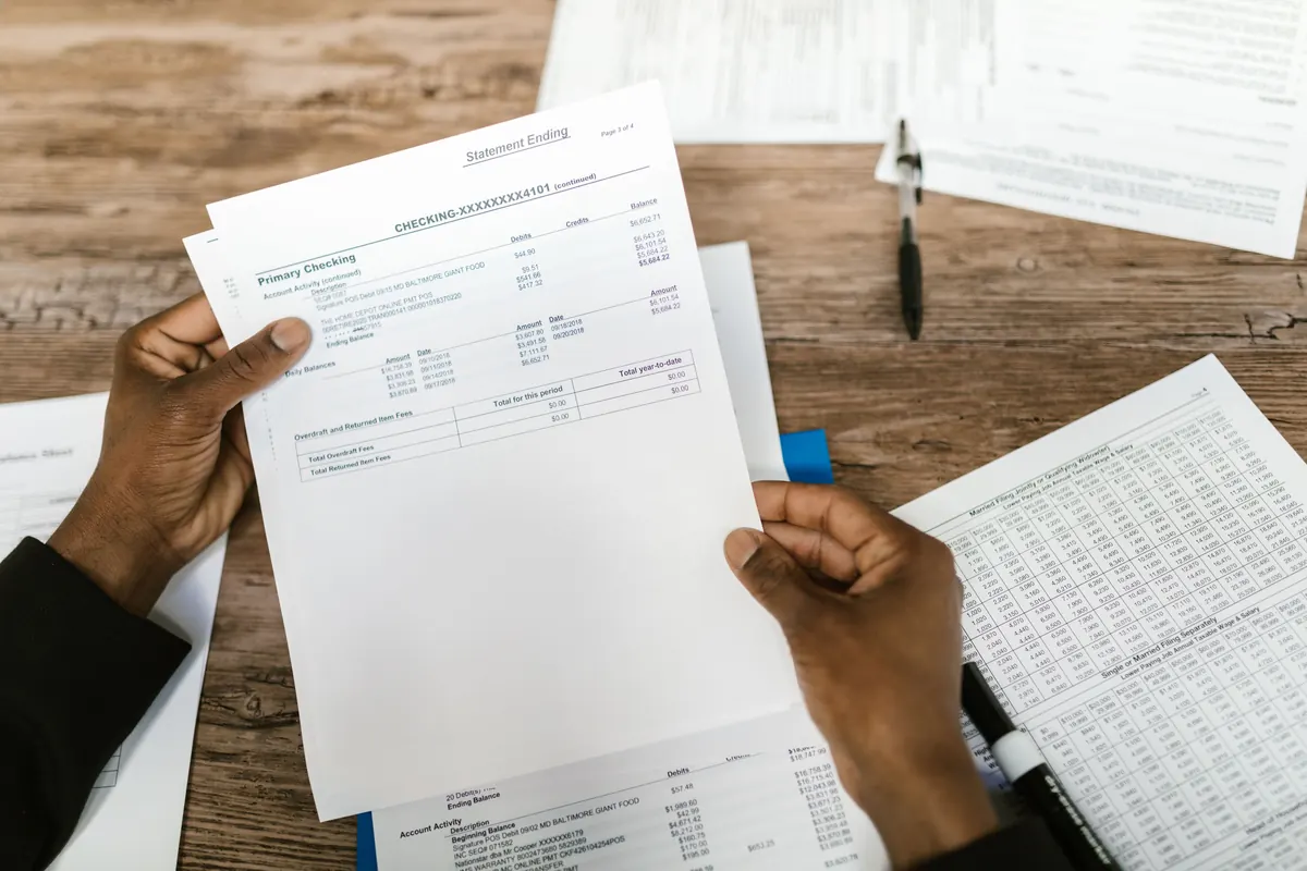 Two hands holding a printed account statement over a wooden desk covered in papers, a pen nearby