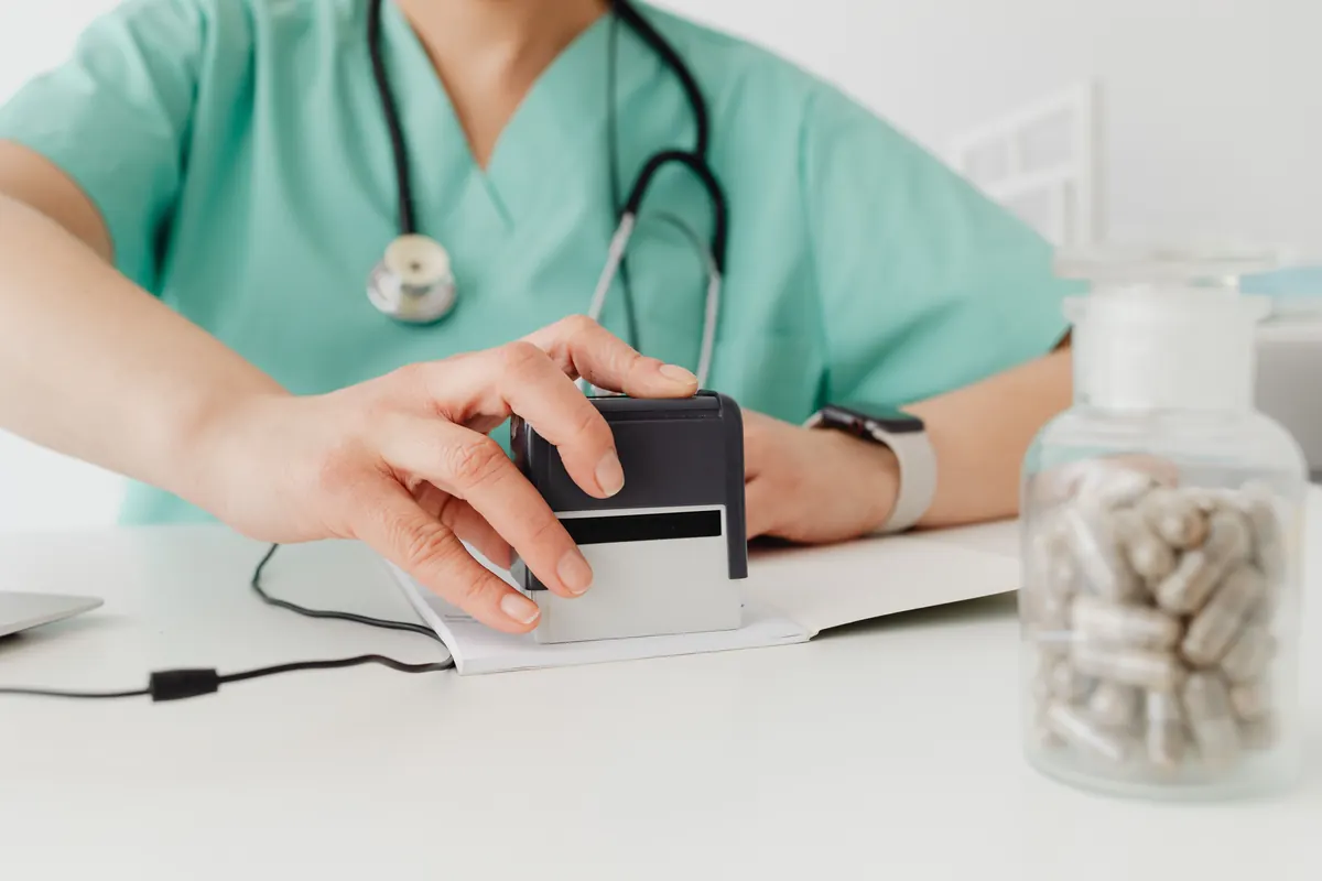 A health worker in green scrubs with a stethoscope pressing a stamp on paper beside a jar of capsules