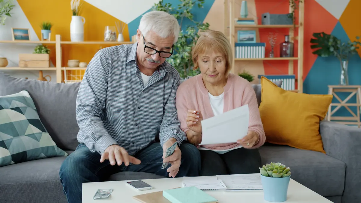 An older couple on a sofa looking over a letter together, with cash, a phone and papers on the table