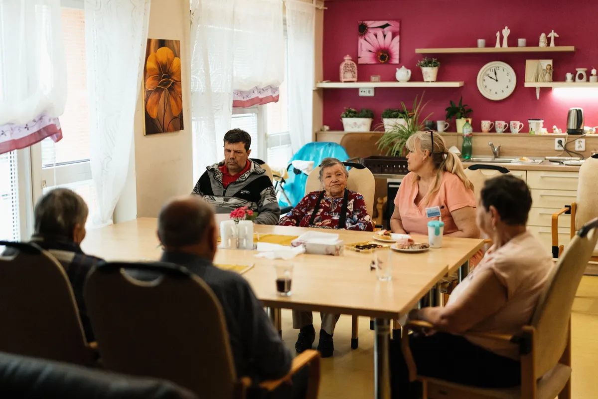 Older residents and a staff member in a pink top sitting around a table in a bright shared dining room