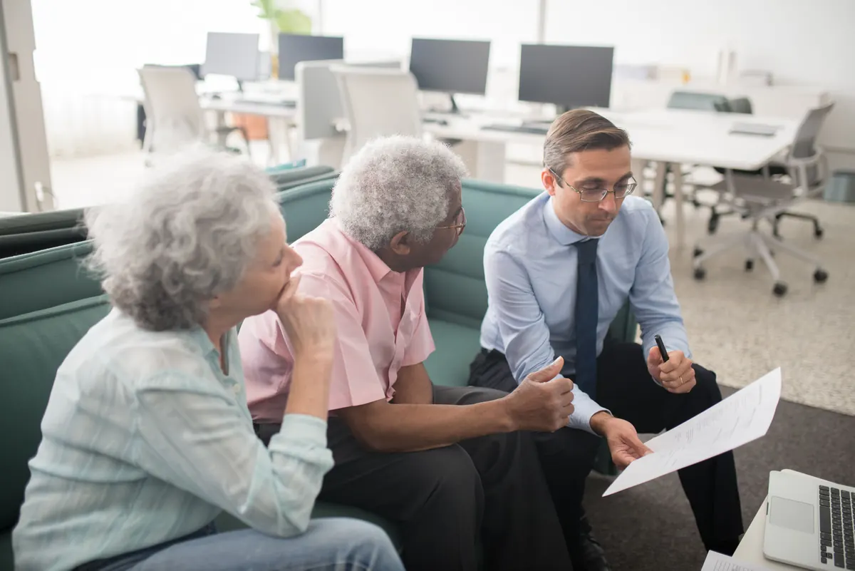 An older couple on a sofa listening as a man in a shirt and tie goes over a printed page with them
