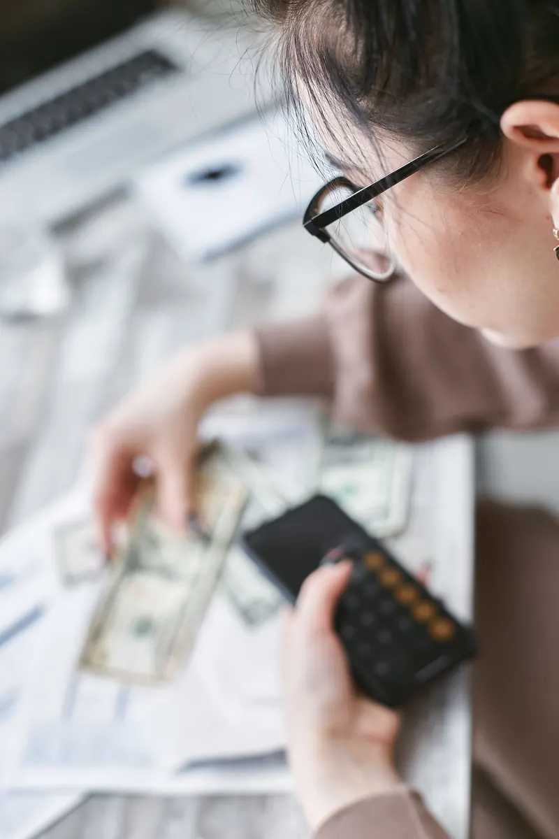 A woman in glasses using a calculator while holding cash over paperwork on a desk