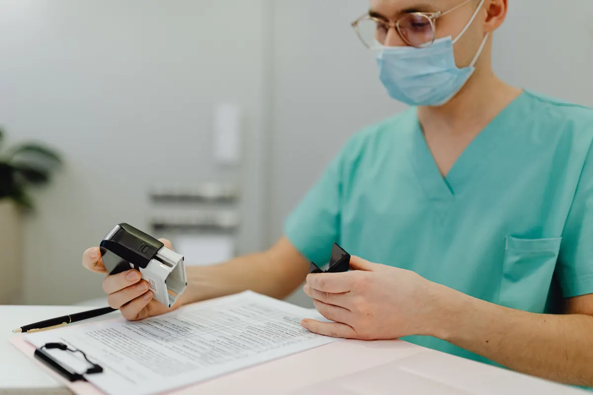 A health worker in green scrubs, a face mask and glasses holding a stamp over a form on a clipboard