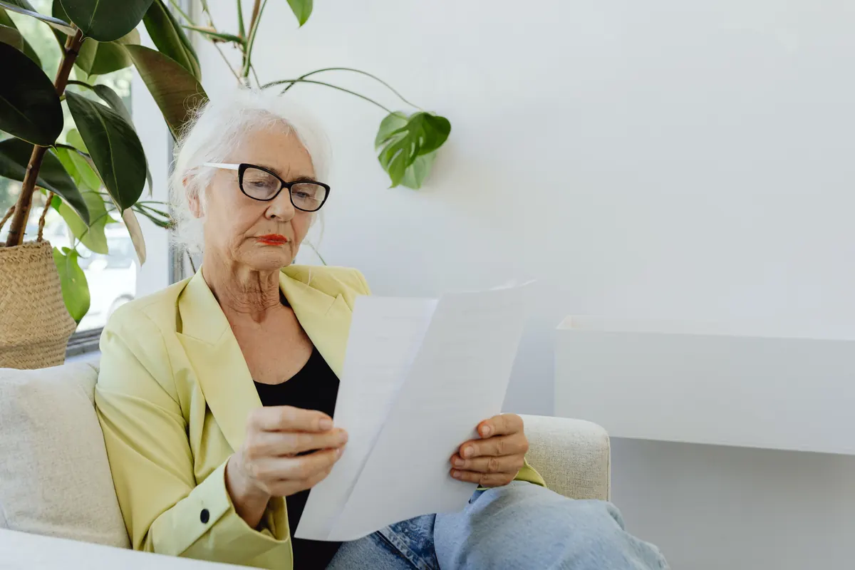 An older woman with white hair and glasses in a yellow blazer reading papers on a sofa