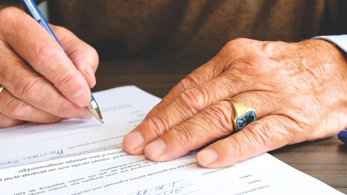 Close-up of an older man's hands signing a printed document with a blue pen, a ring on one finger