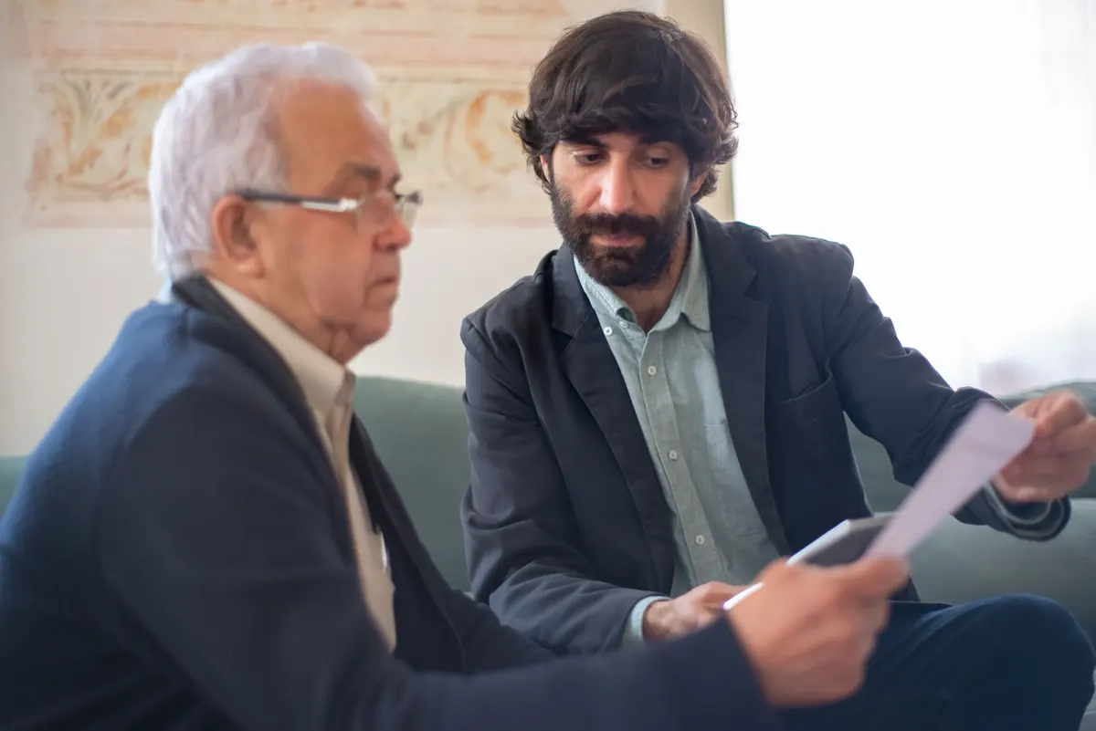 A younger bearded man showing an older man with white hair and glasses a document on a sofa