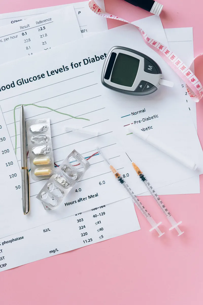 A glucose meter, blister packs of pills, syringes and a pen on printed lab charts on a pink background