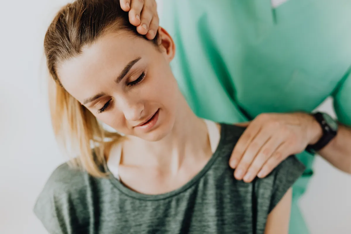 A health worker in green scrubs gently tilting a young woman's head and pressing on her shoulder