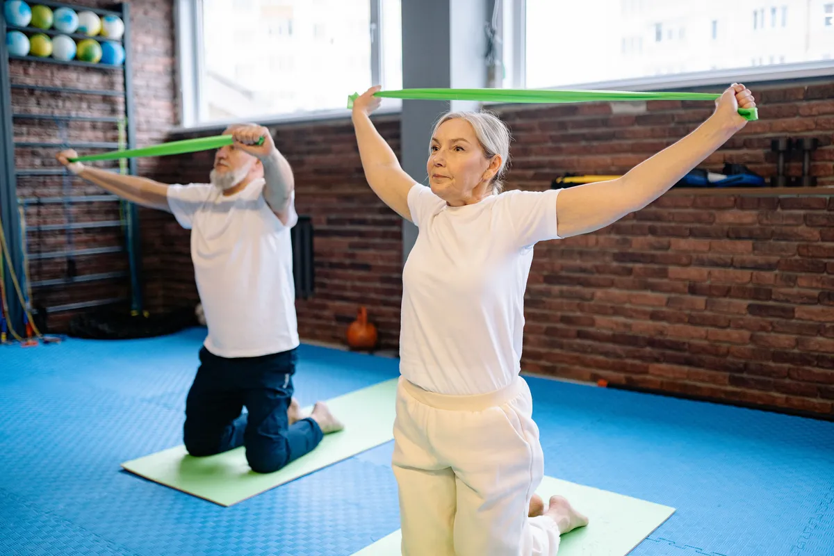 An older woman and an older man kneeling on exercise mats, stretching green resistance bands overhead
