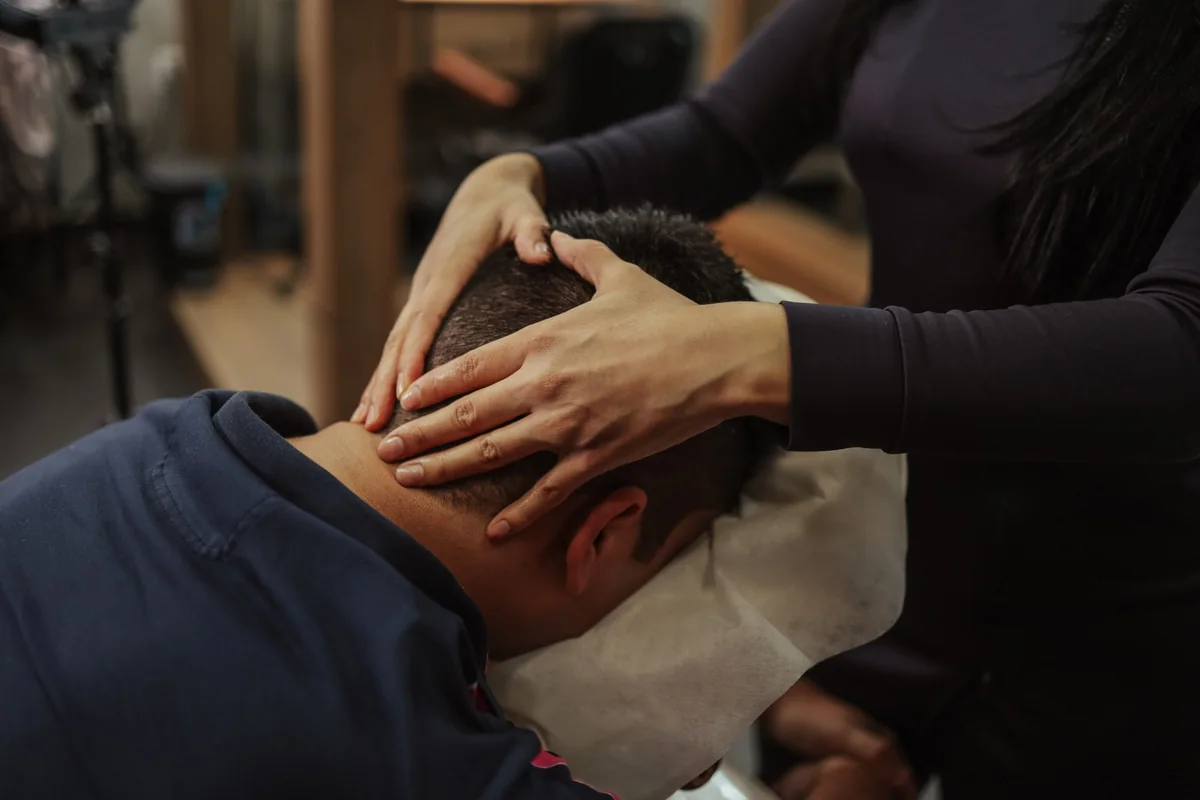 A practitioner's hands resting on the back of a man's head as he leans forward on a massage chair