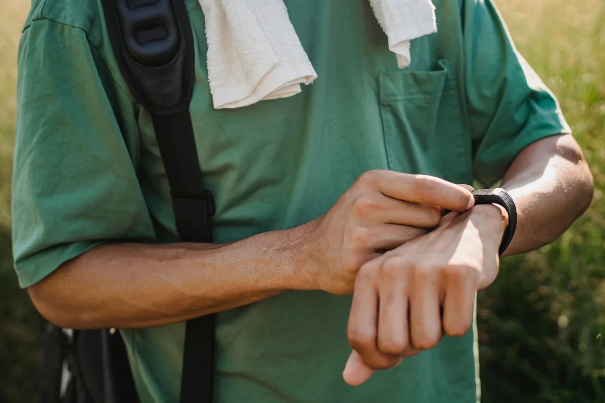 A man in a green T-shirt with a towel on his shoulder checking a fitness band on his wrist outdoors